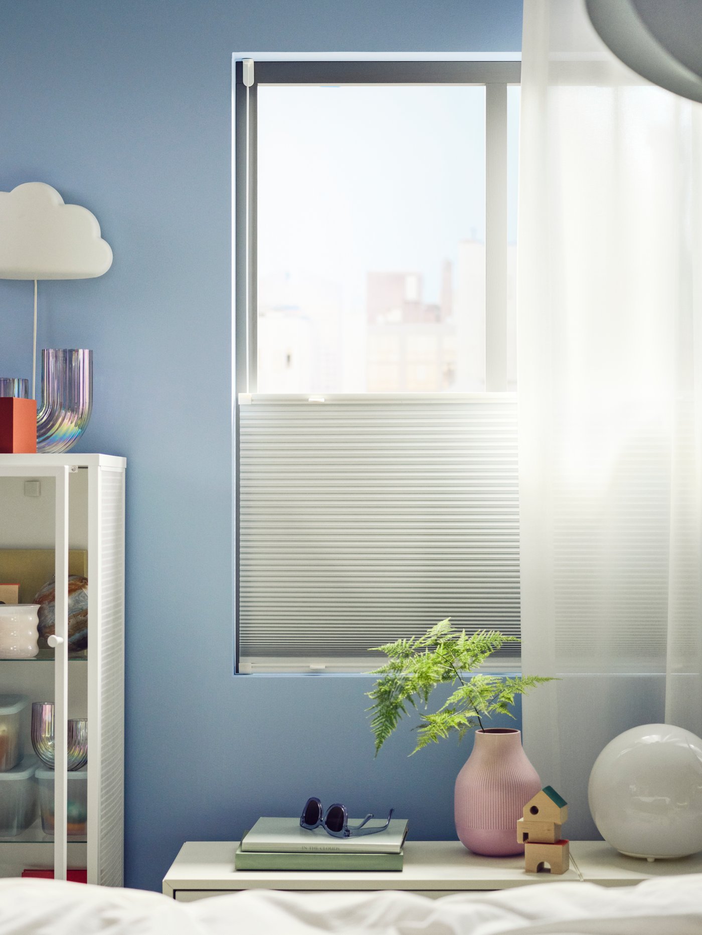 Vor einem lichtdurchfluteten Fenster hängt ein weißes HORNVALLMO Verdunkelungsplissee und ein weißer durchscheinender ROSENROBINA Vorhang. Die Wand ist hellblau gestrichen und neben dem Fenster hängt eine Wandlampe in Form einer kleinen Wolke.
