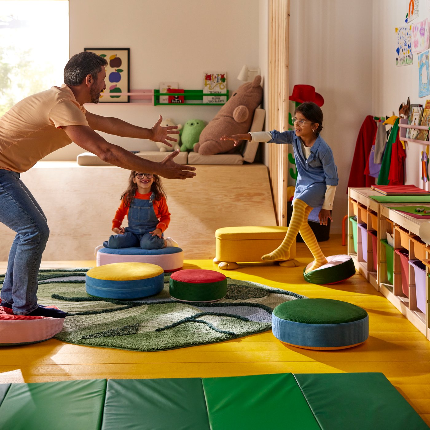 Ein Elternteil und zwei Kinder spielen „Der Boden ist Lava“ mit GREJSIMOJS-Bodenkissen und einem GRESIMOJS-Teppich.