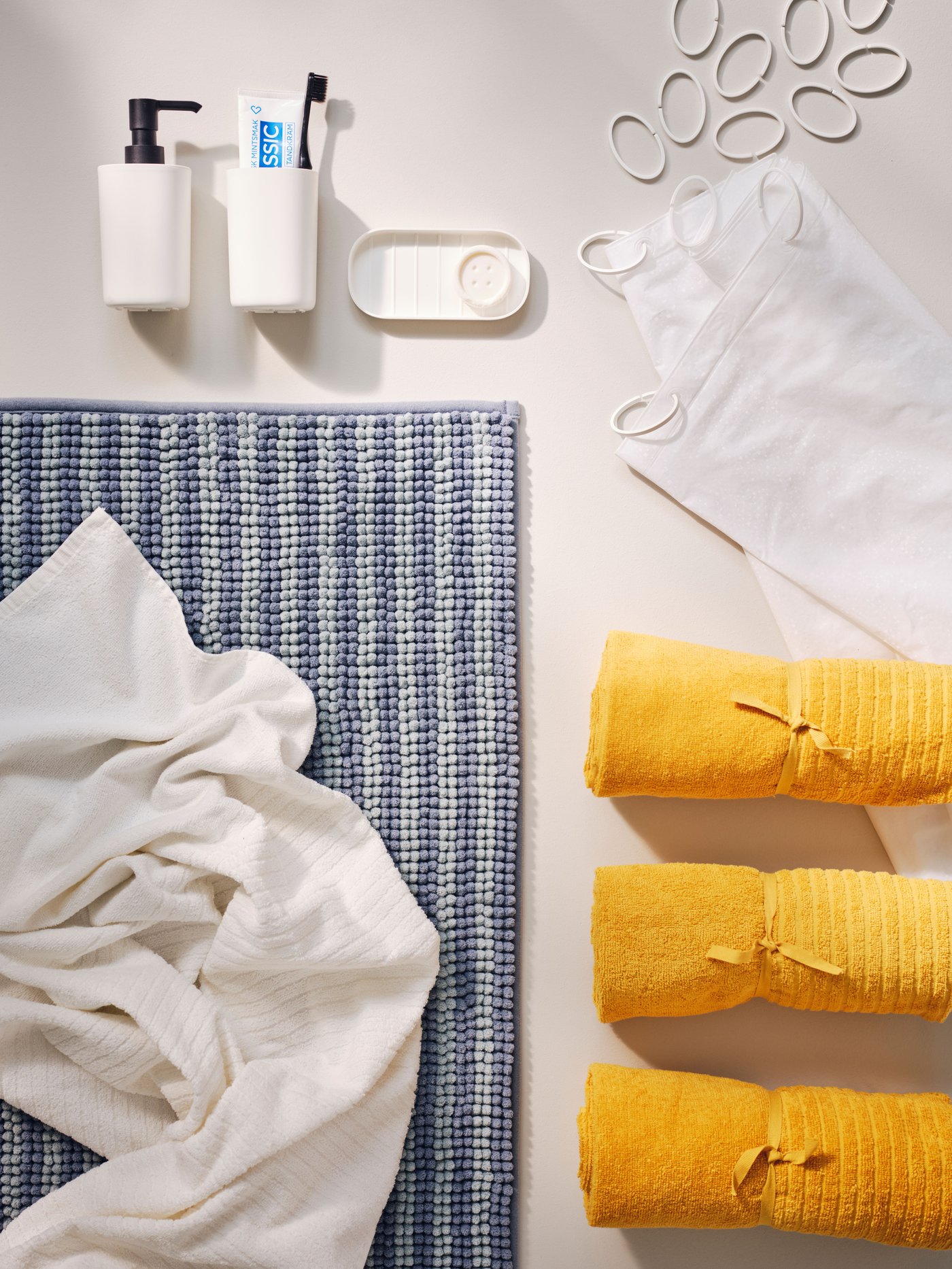 College bathroom starter kit for students with white shower curtain, yellow towel, blue bathmat and white sink accessories.