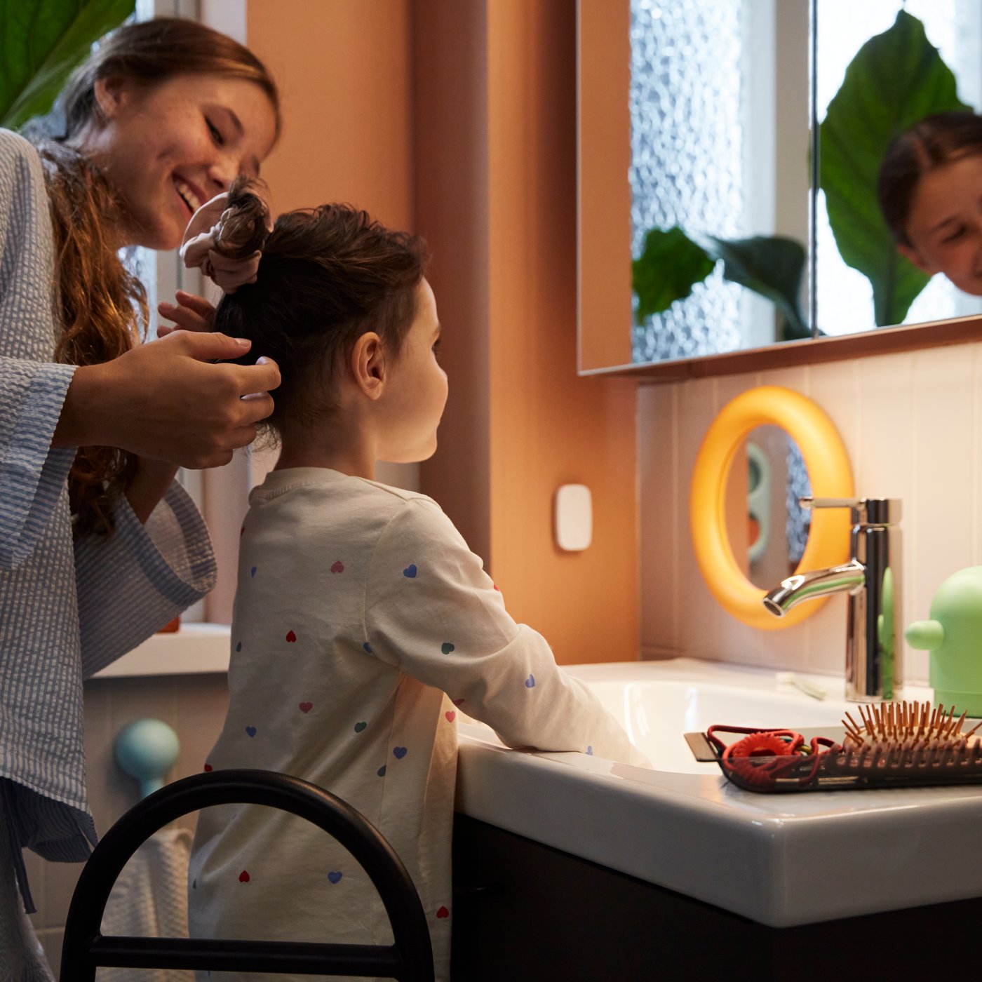 A bathroom with a LETTAN mirror cabinet showing a child getting ready with an adult by a sink with a green plant in the back.