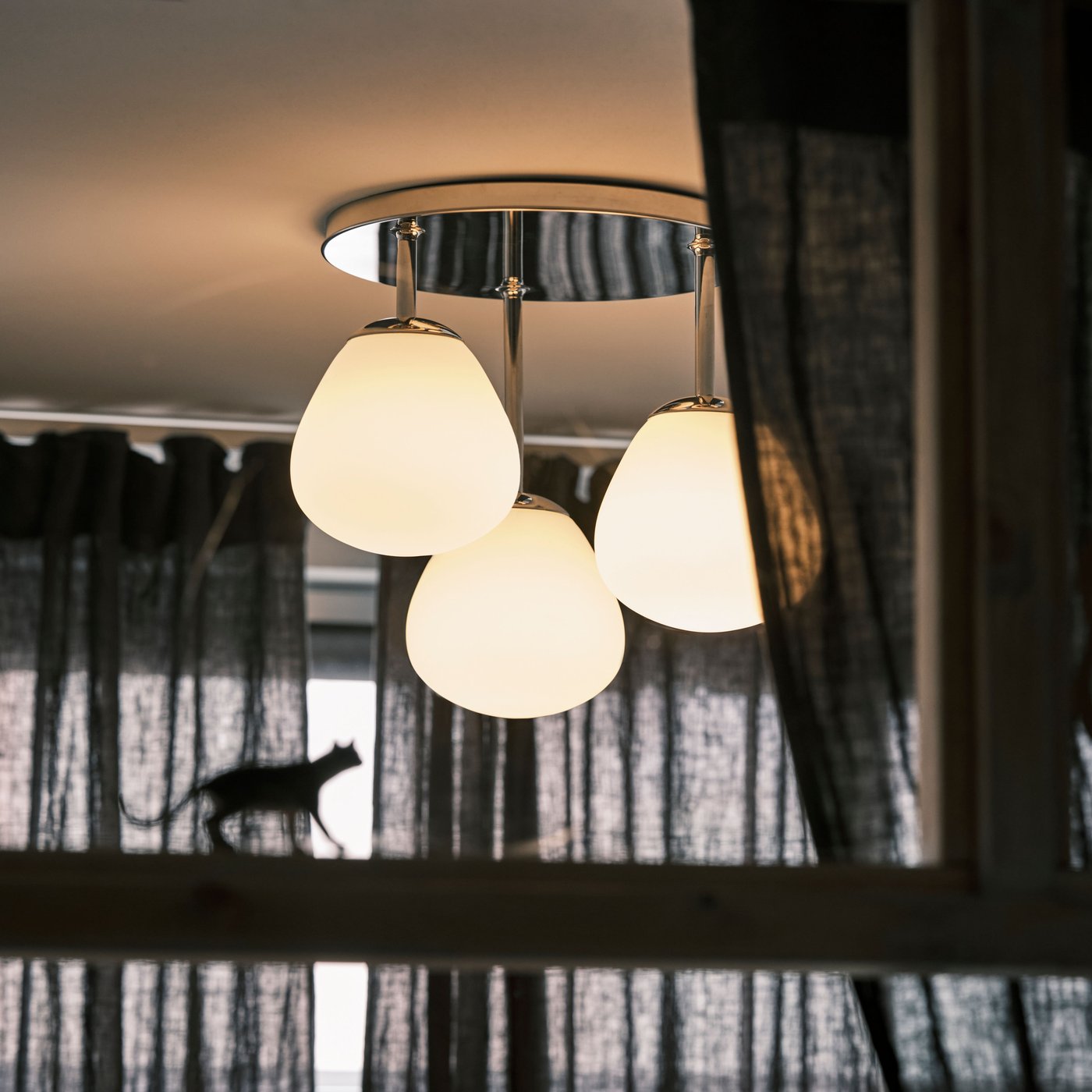 A DEJSA chrome-plated ceiling lamp with three opal white glass lamps is placed in front of a window with dark sheer curtains.