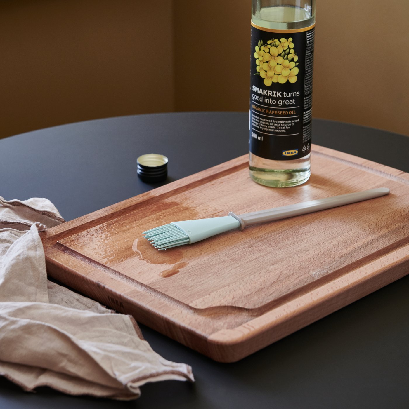 A PROPPMÄTT chopping board in beach with a small brush and a bottle of rapeseed oil on a table beside a cloth.