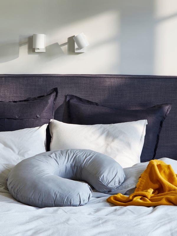A grey LEN nursing pillow sits on top of a DUNVIK divan bed along with a dark yellow SOLGUL blanket in a bedroom.