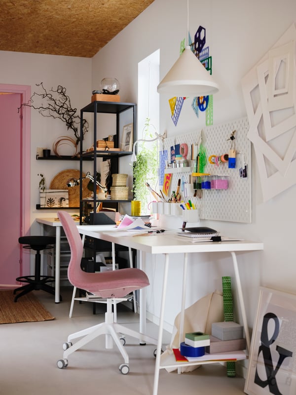 A colorfully decorated workspace with a white LAGKAPTEN tabletop, white SKÅDIS pegboards and a pink LÅNGFJÄLL swivel chair.
