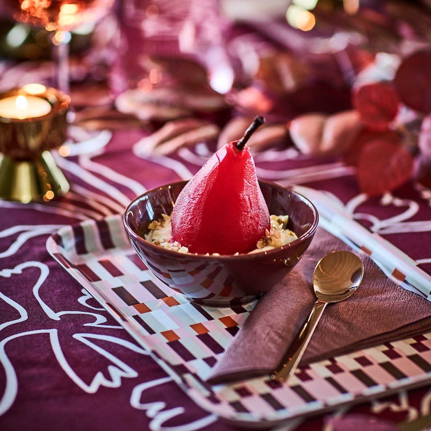 Un dessert con una pera cotta rosa in una ciotola HÖSTAGILLE e un tovagliolo di carta marrone-rosso piegato su un vassoio HÖSTAGILLE con un motivo colorato.