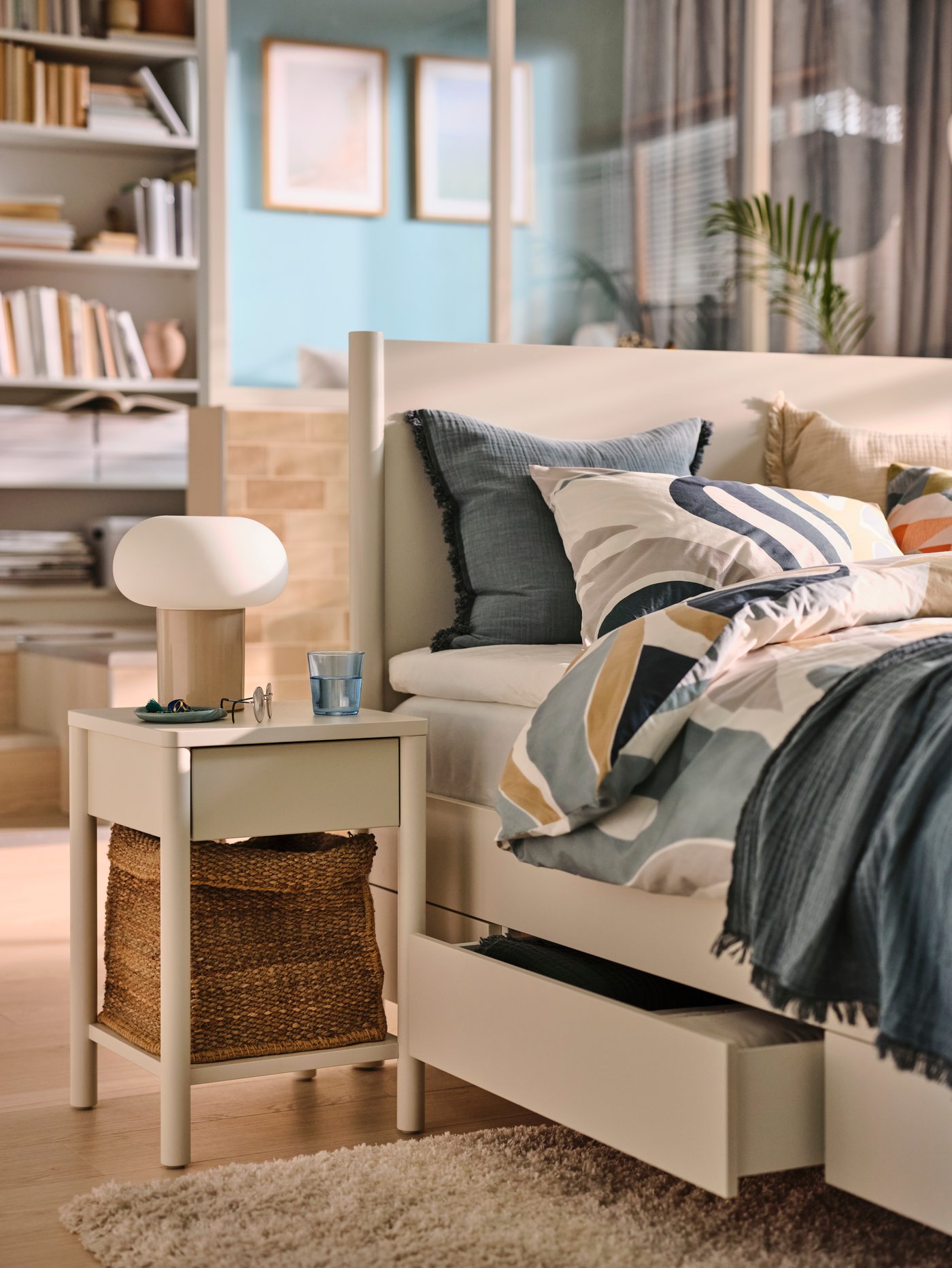 An off-white TONSTAD bedside table beside an off-white TONSTAD bed covered in MYGGLASVINGE multicolour bed linen.