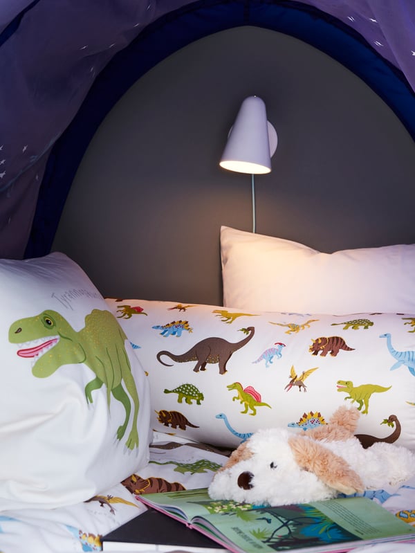 Inside a KURA bed tent mounted on a KURA reversible bed with JÄTTELIK dinosaur bed linen and cushion, and a wall lamp.