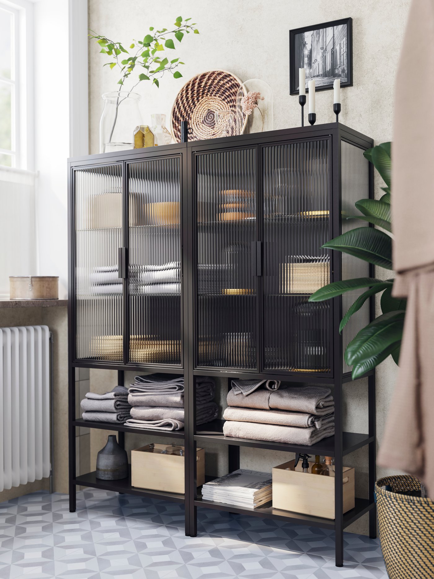 Es ist ein graues Badezimmer mit schwarzen MOSSJÖN Vitrinen mit Badezimmerutensilien auf Böden, gefalteten Handtüchern, Boxen und Deko zu sehen.