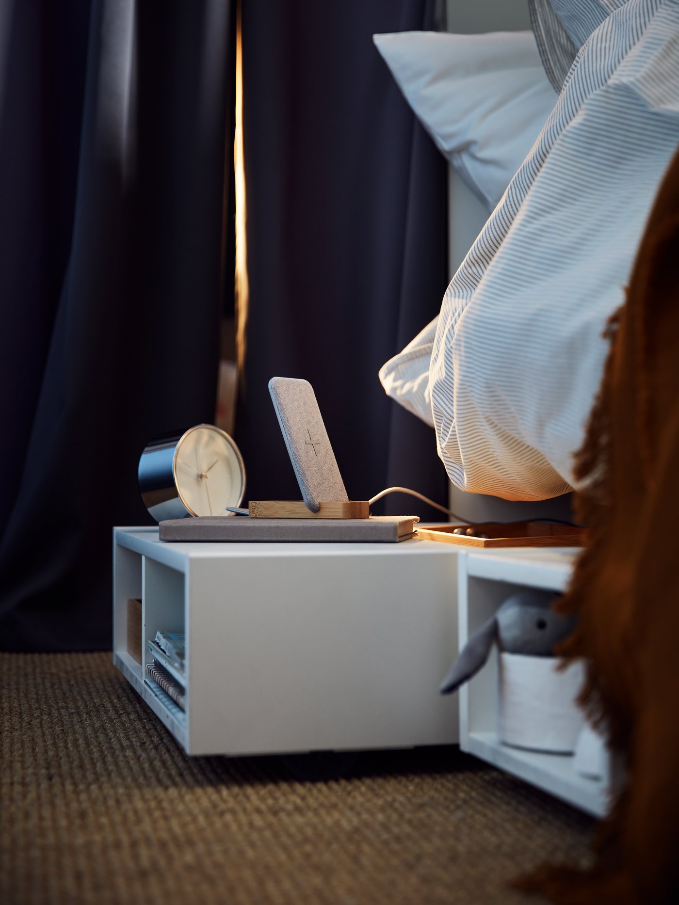 Two white FREDVANG underbed storage units/bedside tables holding books and various other items, on a jute rug under a bed.