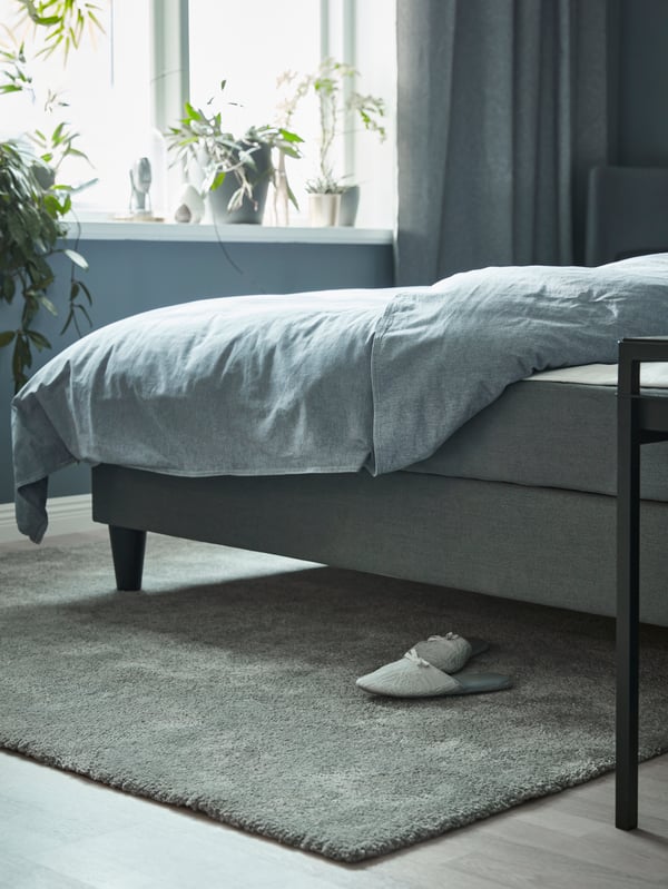 A SÄBÖVIK divan bed with KOPPARBLAD bed linen stands on top of a STOENSE rug with a pair of slippers near a window.