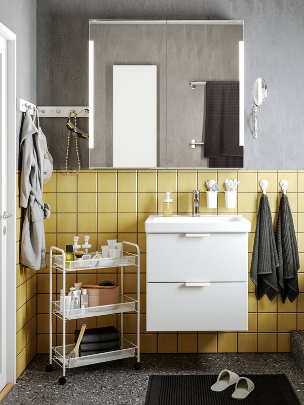 Un baño con cerámicas amarillas. Destaca el mueble del lavamanos GODMORGO blanco y al costado un carrito de baño HORNAVAN.