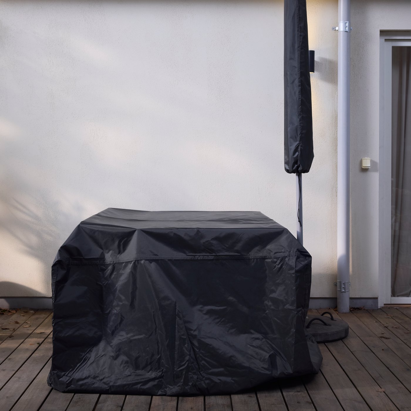 A TOSTERÖ cover for outdoor furniture set covers a piece of furniture next to a parasol covered by a TOSTERÖ parasol cover.