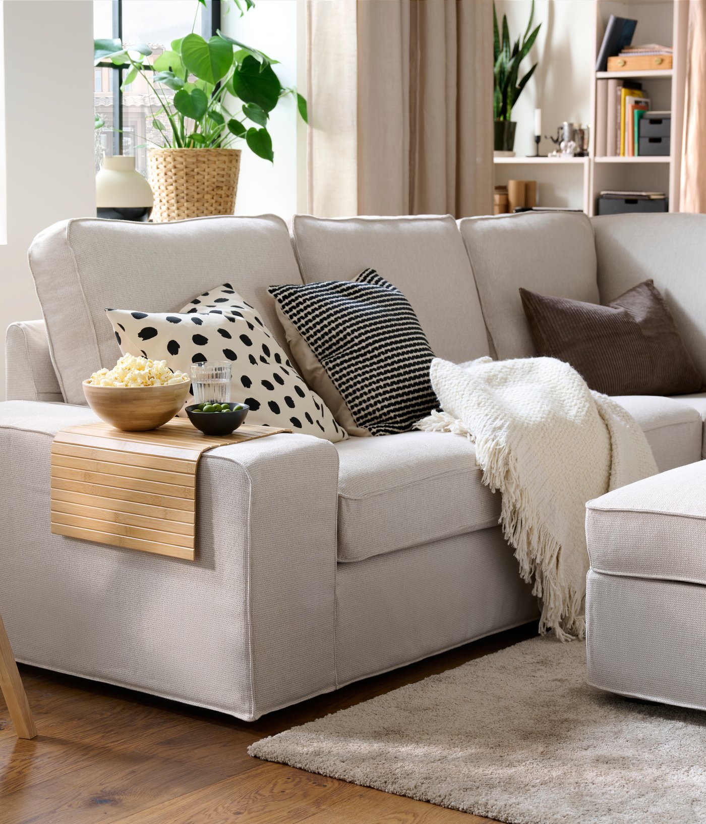 A light living room with a KIVIK corner sofa with colourful cushions, a throw, and a RÖDEBY armrest tray holding snack bowls.