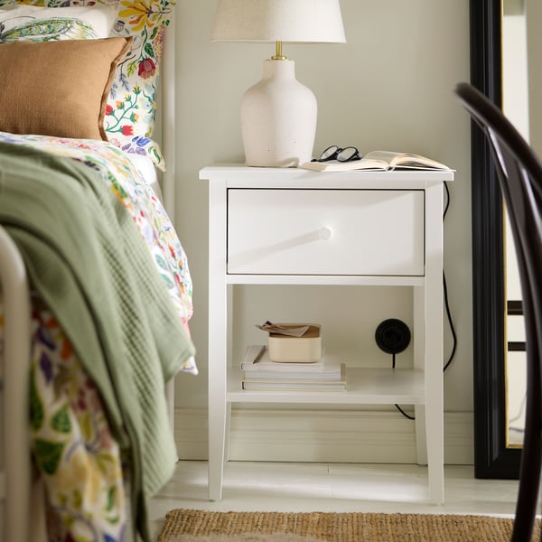 A white GULLABERG bedside table with an off-white BILDVÄDER table lamp on it beside a bed with floral bed linen on it.