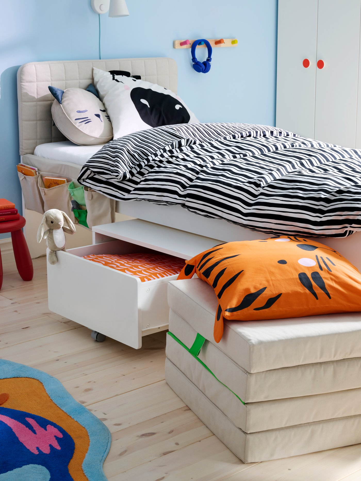 EIn SLÄKT Kinderbett mit Kopfteil, geöffneter Schublade und einer faltbaren Matratze. Auf dem Bett liegt eine Decke mit gestreifter NATTHÄGER Bettwäsche.