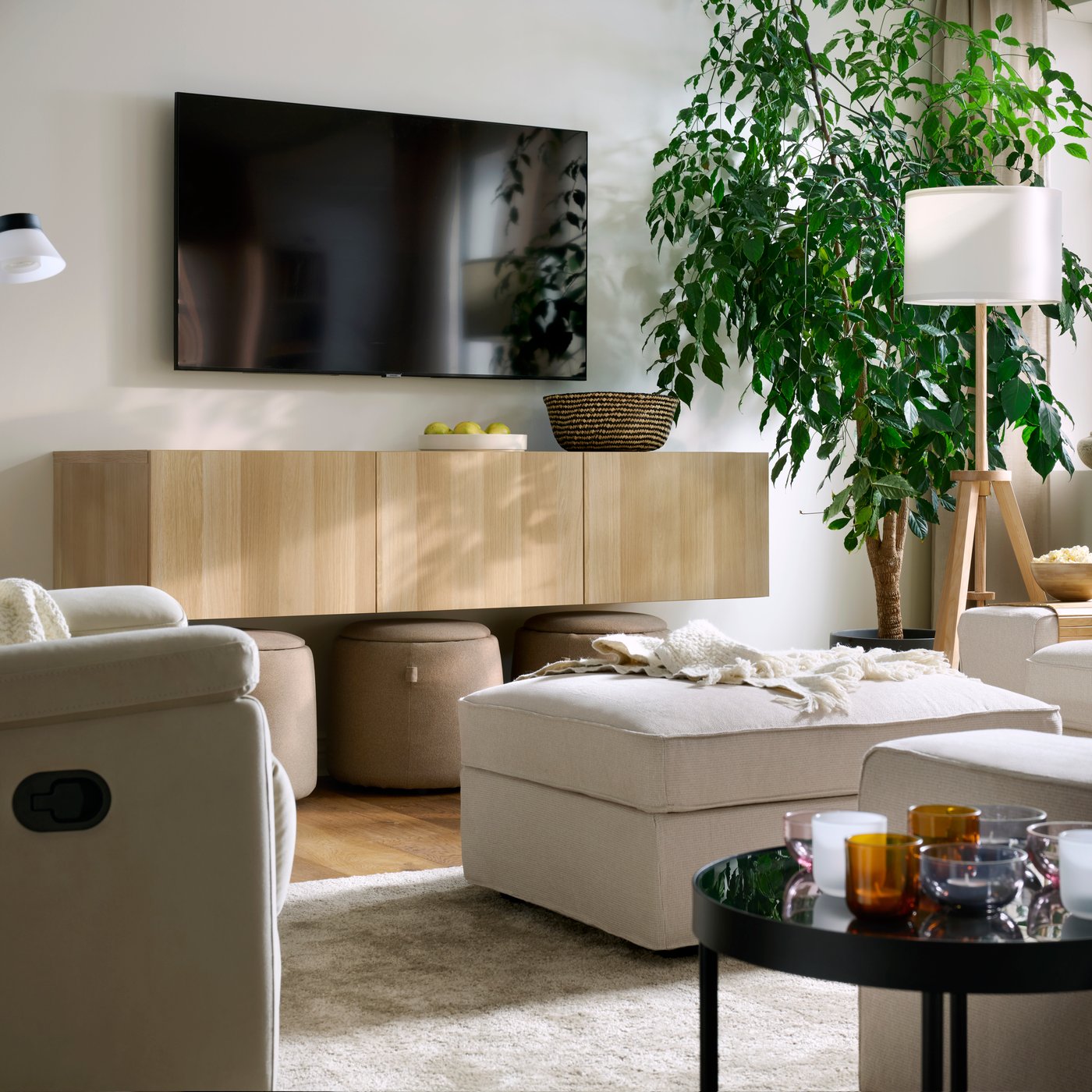 A light, bright living room with a KIVK pouffe, a SKULNÄS recliner, a STOENSE rug, a wall-mounted TV and a big, leafy plant.