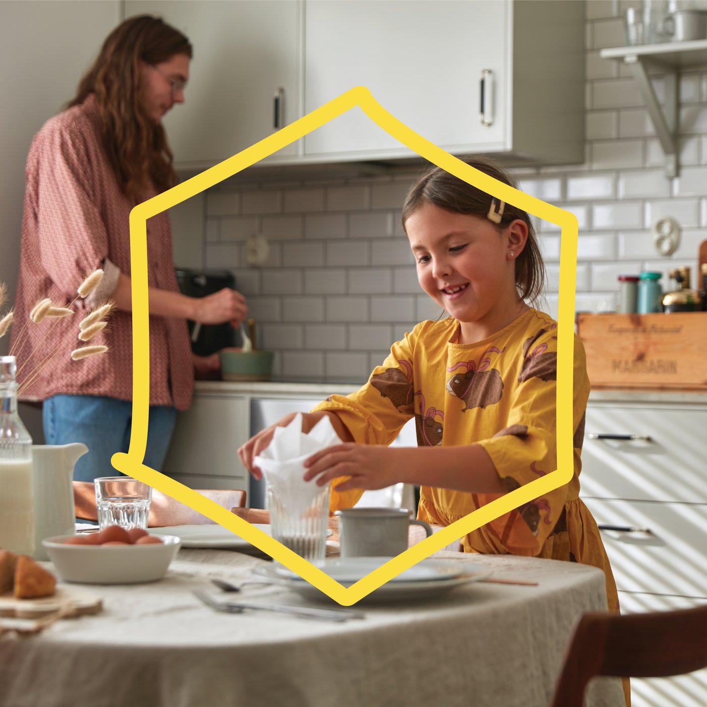 A child in a yellow dress places a napkin into a glass at a table, while another person stirs ingredients in a bowl in a bright kitchen with white subway tiles.