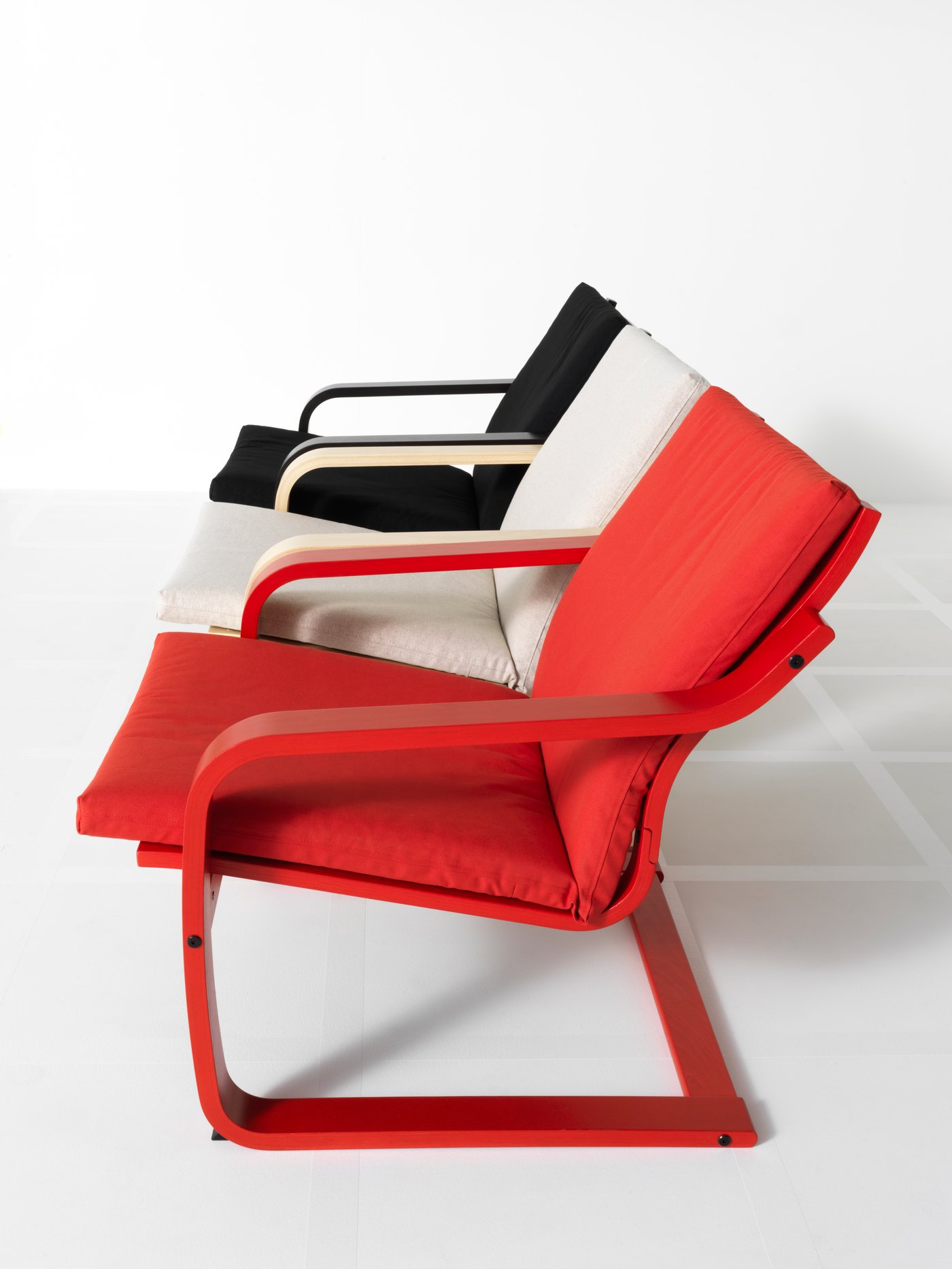 Three POÄNG low-back armchairs in red, beige/birch and black stand on a white floor in a straight vertical line.