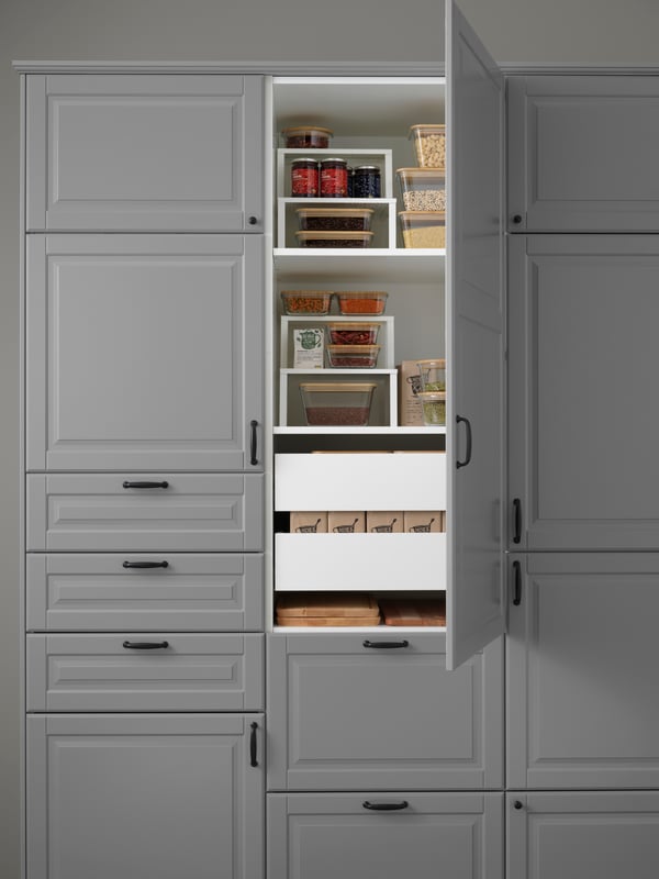 Close-up of kitchen cabinet with grey doors, white drawers and shelves, food containers with lids, chopping boards, jam jars.