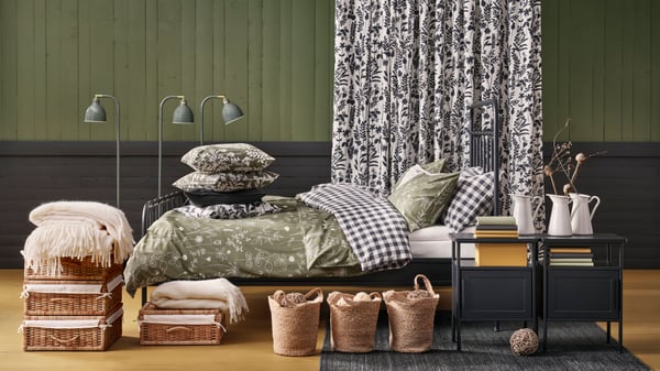 An anthracite STJÄRNÖ bed dressed with SLÅNGULDMAL and EKTANDVINGE duvet covers. Rattan boxes and jute bags stands in front.