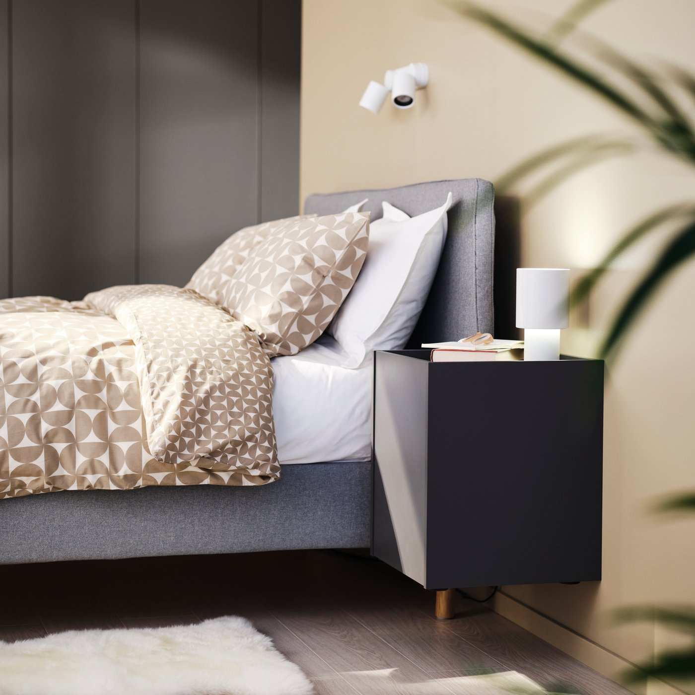 A dark grey PLÖJKER bedside table attached to the wall beside a beige-grey TÄRNKULLEN bed with ÄNGSNEJLIKA bed linen on it.