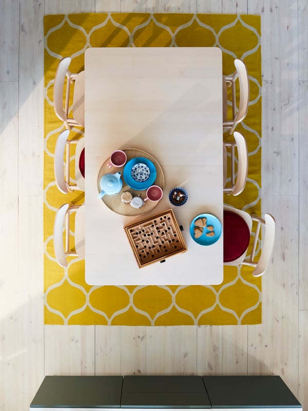A teapot, cups, biscuits and a board game on a table in birch, six chairs in birch, two red chair pads and a yellow rug.