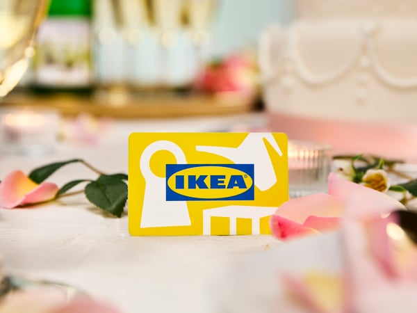 Carte cadeau IKEA jaune posée sur une nappe blanche devant une rose rose et un petit bougeoir en verre