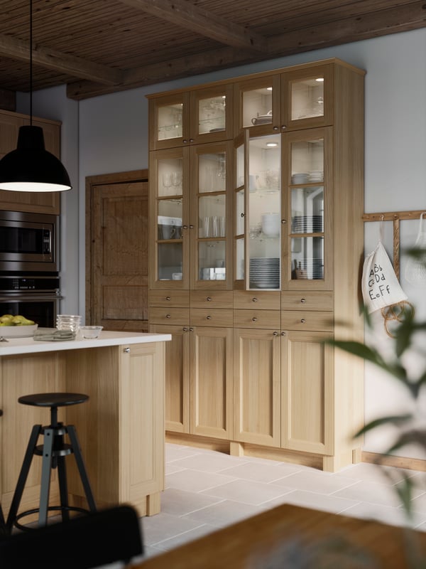 Two high cabinets in a kitchen with closed storage below and VEDHAMN glass doors on the top with integrated lighting.