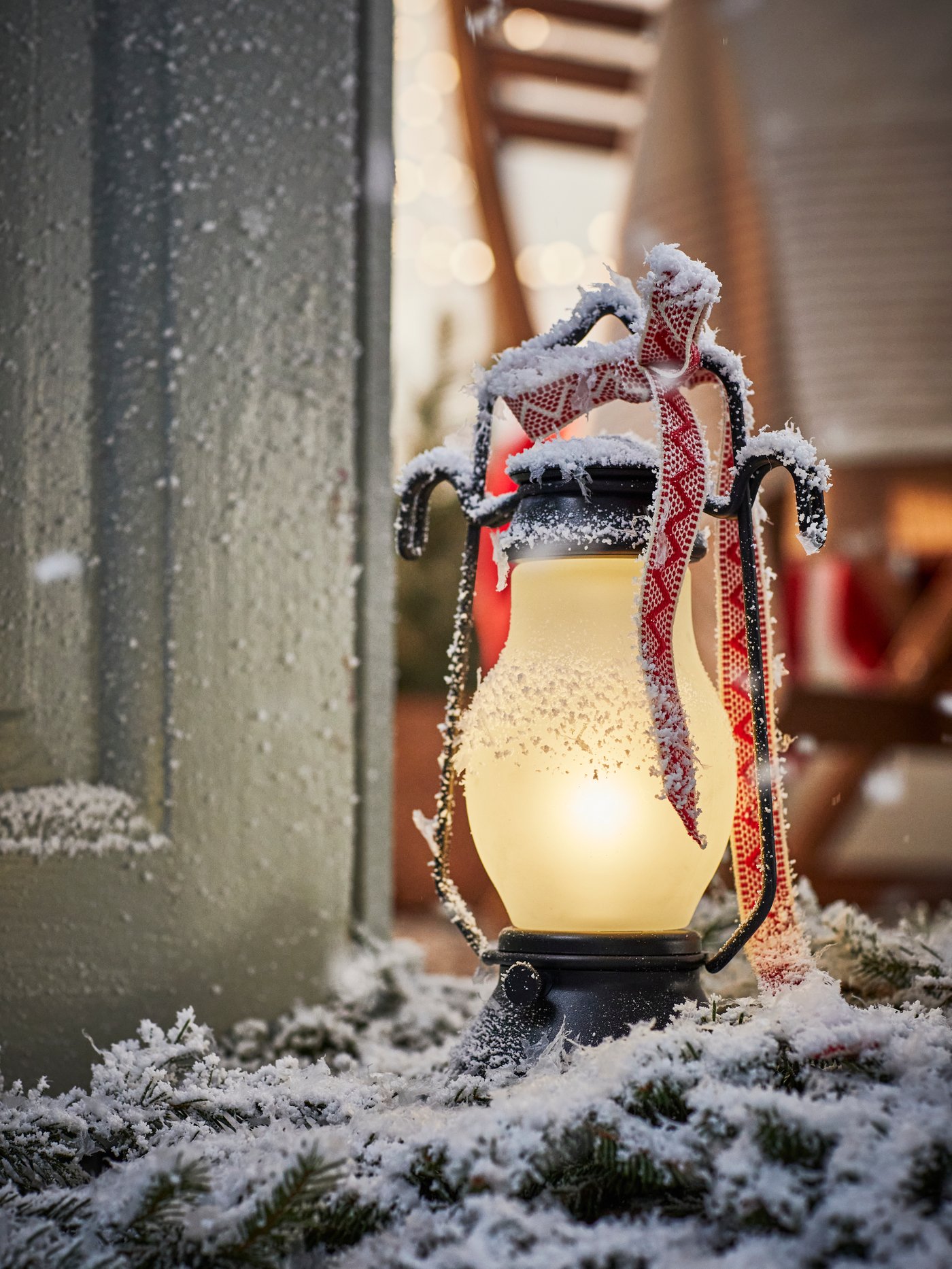 Battery operated STRÅLA LED table lamp with festive red ribbon in snow beside a door at Christmas time.