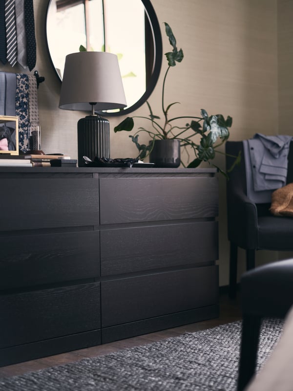 A black chest of drawers with a plant and SNÖBYAR table lamp on top. A rail of ties and a round mirror are on the wall.