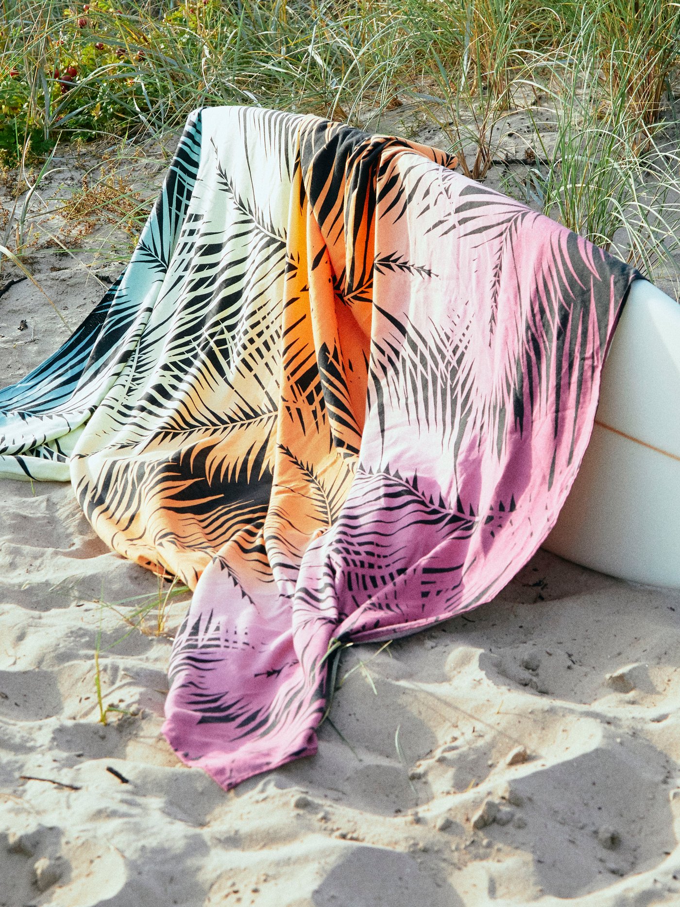 A colourful KÅSEBERGA beach towel with a palm-inspired print displayed across a white surfboard on a sandy beach.