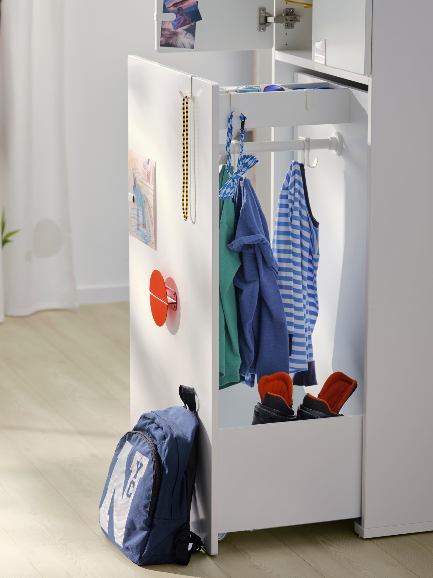 A white SMÅSTAD wardrobe with pull-out unit in a child’s room with clothes on the hanging rail and a bag leaning against it.