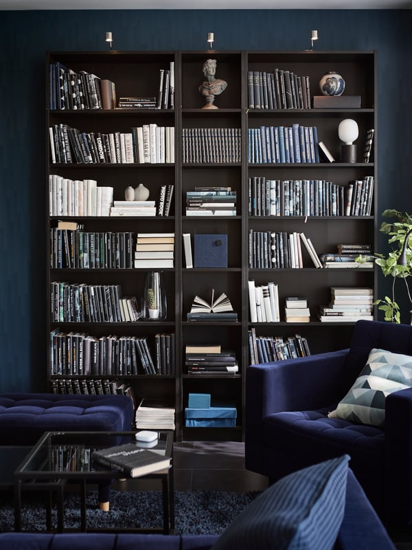 A black-brown BILLY bookcase in a blue living room filled with lots of books and a few objects. A blue armchair is in front.