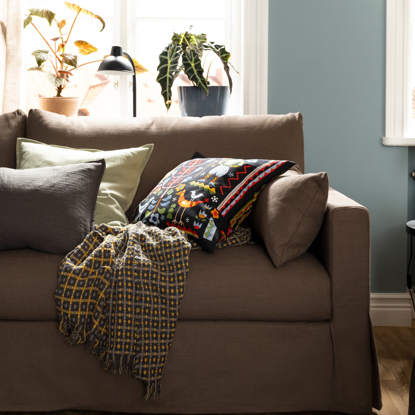 A FLIKBRUNÖRT throw and some cushions, including one with a multicolour TRATTKAKTUS cushion cover, lie on a HYLTARP sofa.