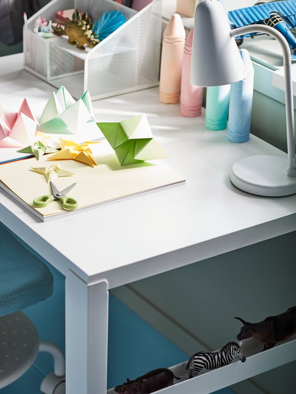 Un escritorio PAHL blanco con útiles de escritorio, juegos de manualidades y origami, y una lámpara de escritorio FUBBLA blanca. 