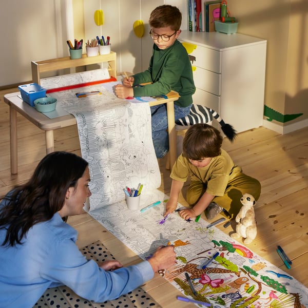 Two children and an adult are in a sunlit children’s room painting on a SANDLÖPARE colouring roll.