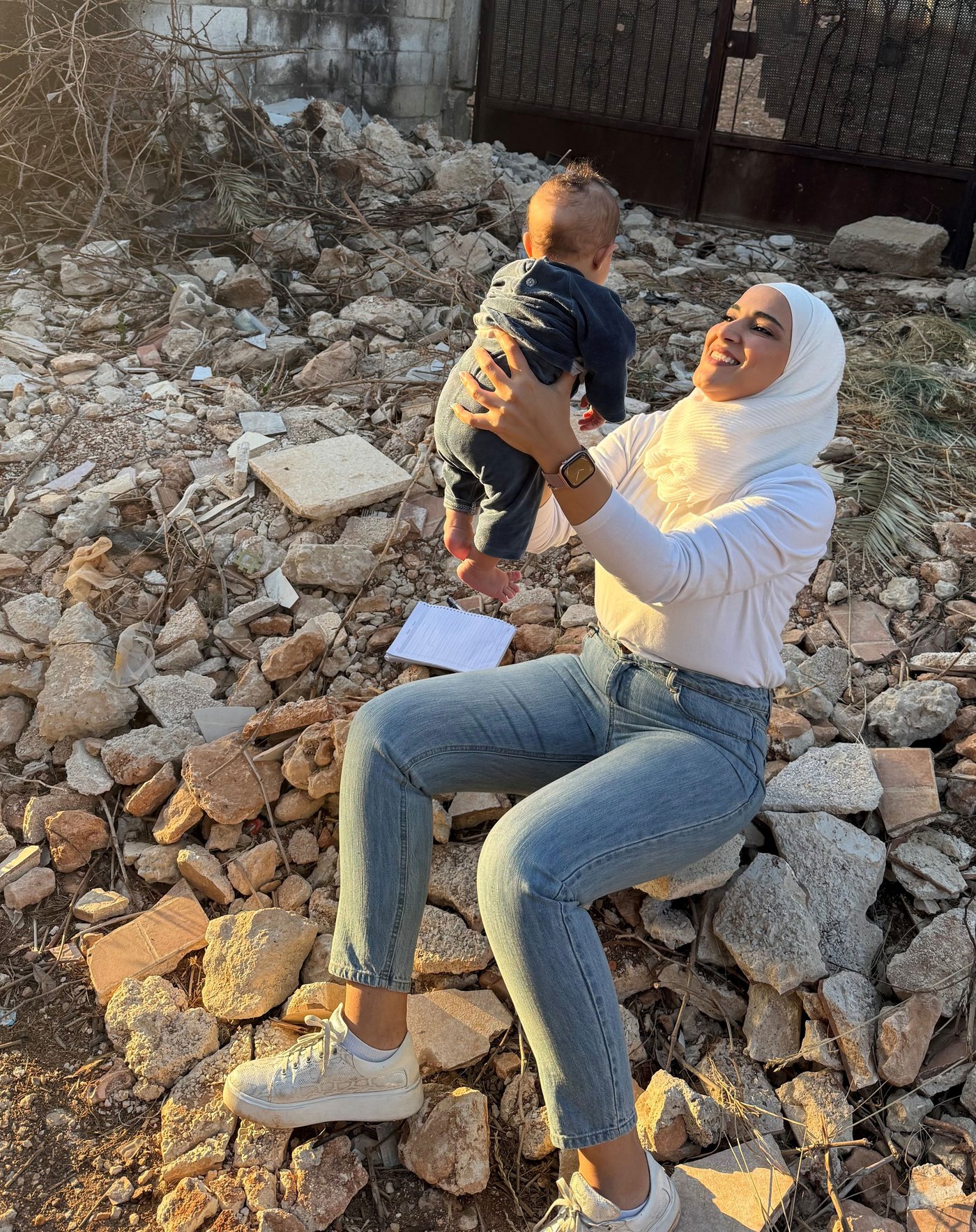Lujain sitzt draussen auf einem Felsen und hält ein Baby in den Armen. Sie lächelt.
