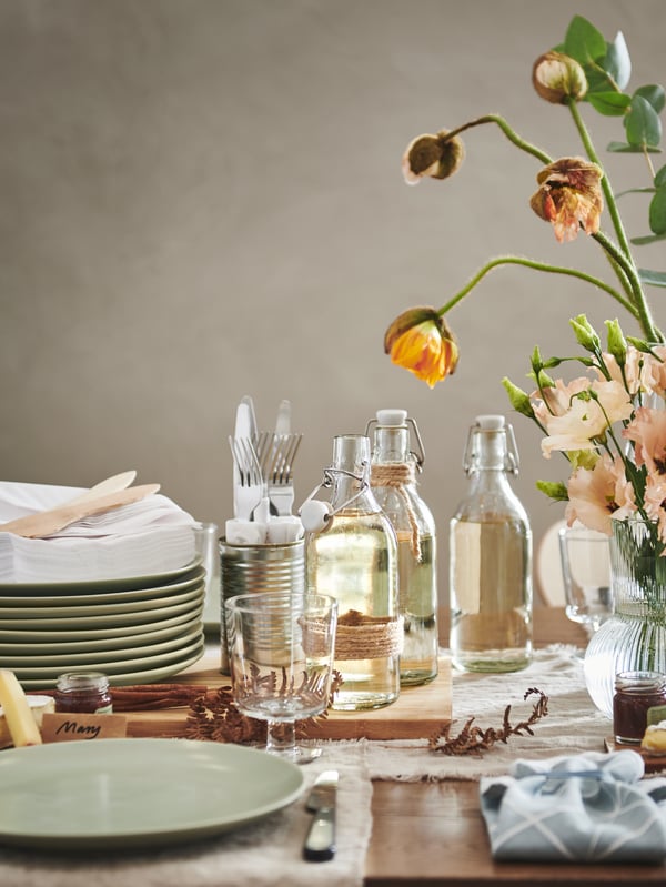 A section of a festively set table with stacked matt-green FÄRGKLAR plates, KORKEN bottles, IKEA 365+ goblets, and flowers.