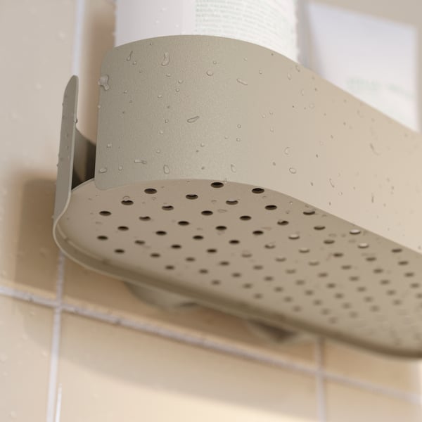 A close-up, underside view of a grey-beige FLORSJÖN shower shelf on a tiled wall, showing its small drainage holes.
