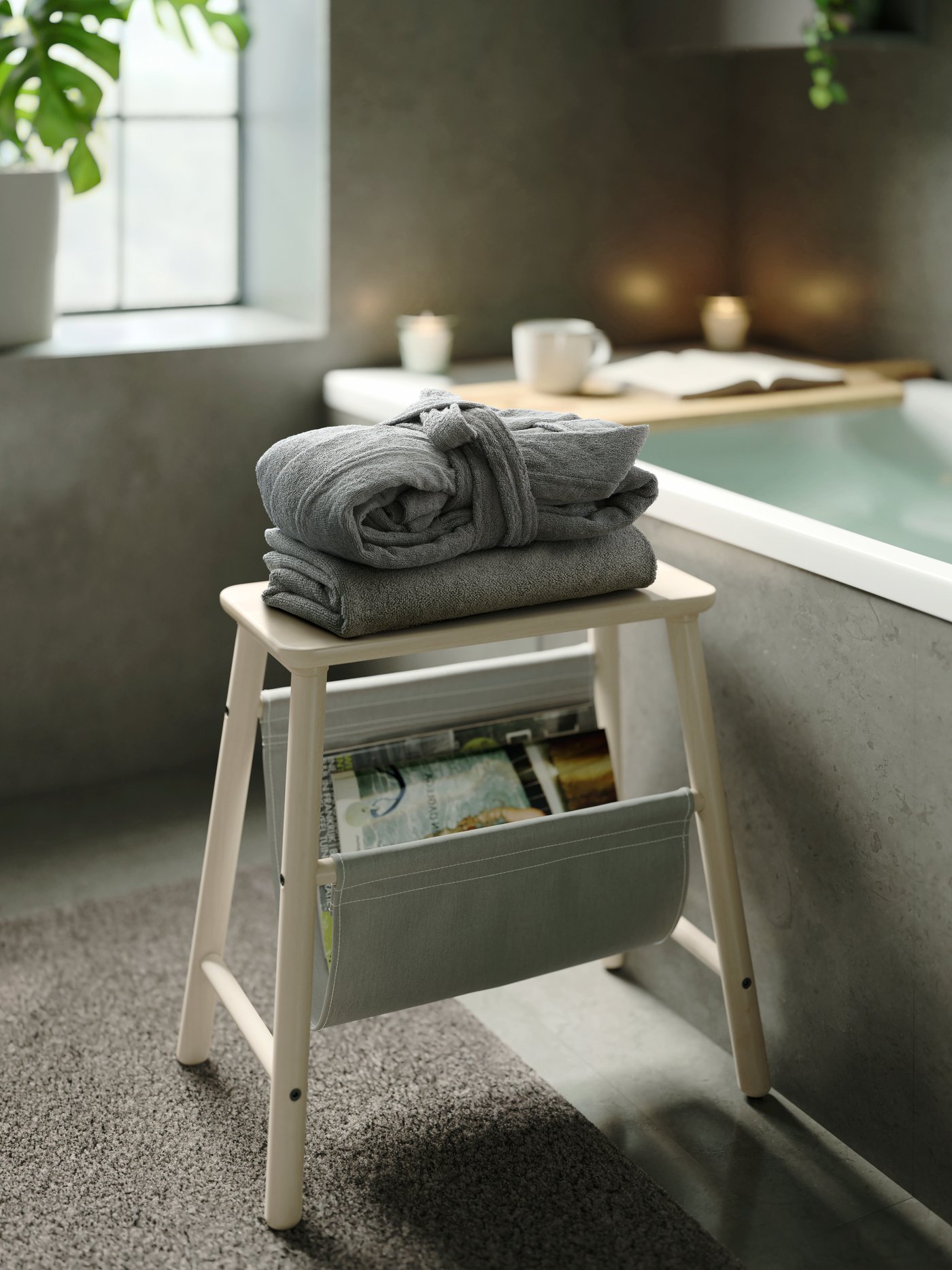 Grauer ROCKÅN Bademantel auf einem Holzhocker neben der Badewanne, auf deren Rand ein Tablett ruht.