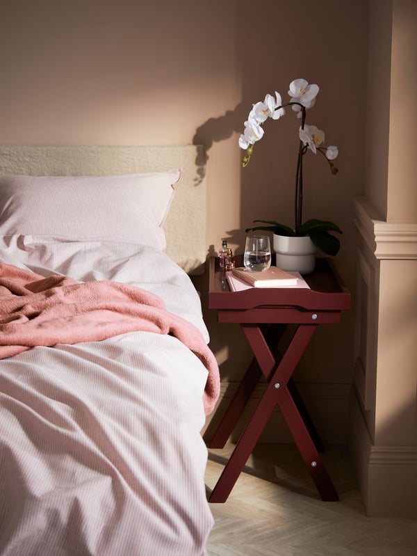 In the corner of a bedroom a tray is placed beside a bed. On the tray are a glass of water, plant pot with an artificial plant, and books.