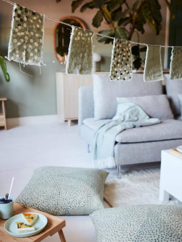 A textile garland in green hanging in a living room with green cushions and a bamboo tray on the floor below.