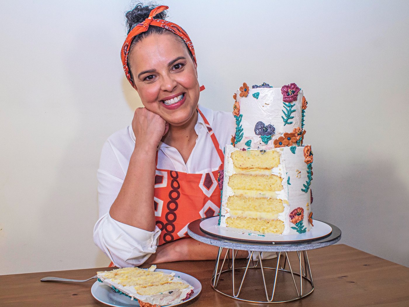 Nasmejana žena u kecelji sedi za stolom, pored visoke, lepo ukrašene torte.