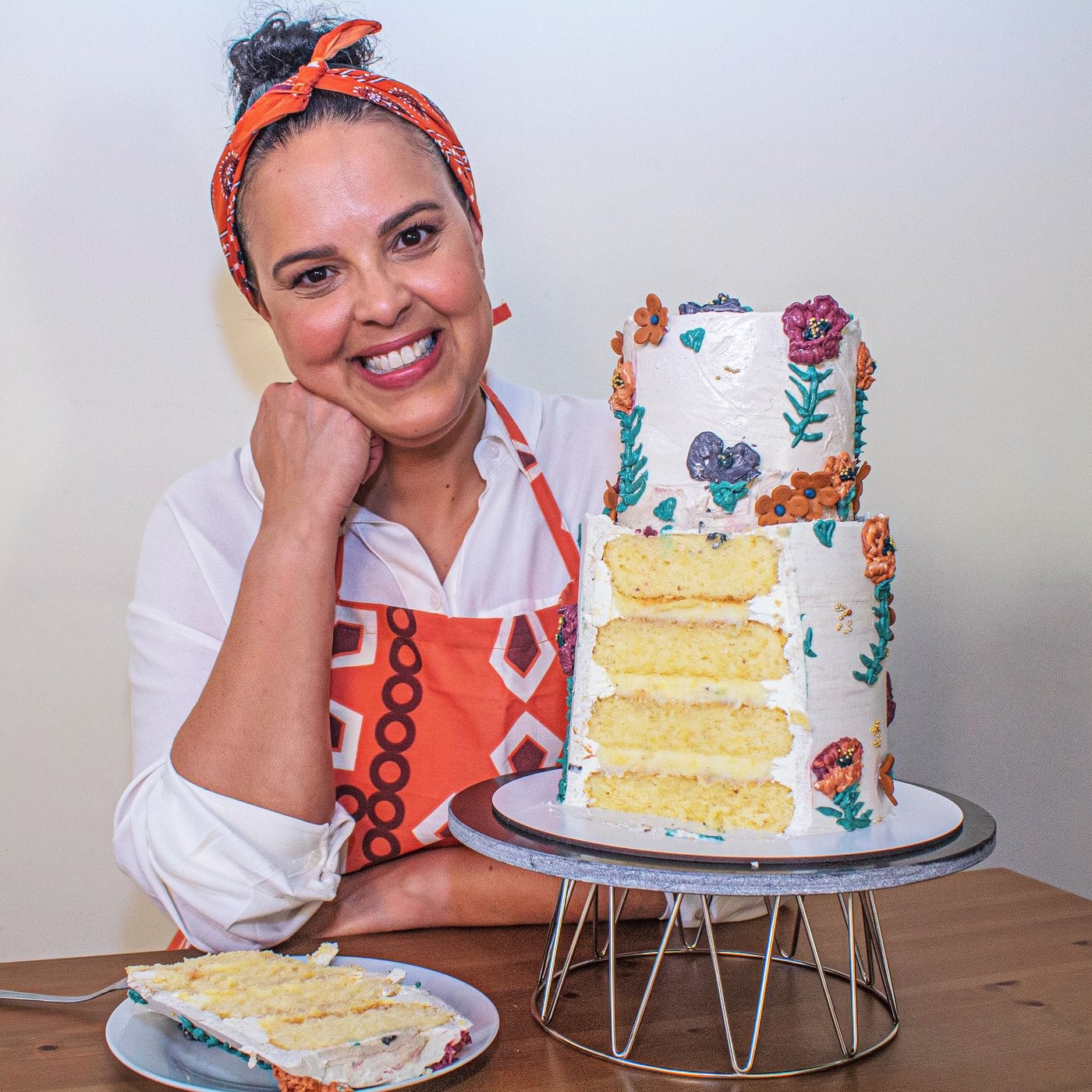 A smiling woman in an apron is sitting at a table, next to a tall, beautifully decorated layer cake.