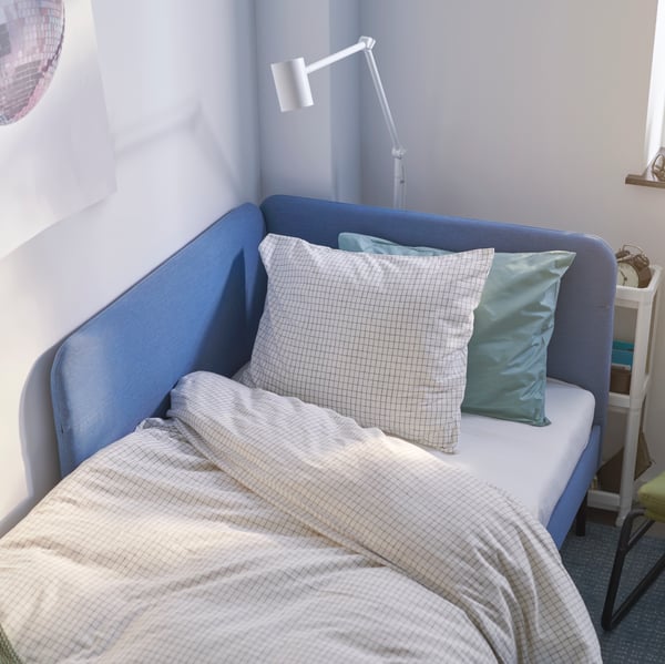 Katil upholsteri BLÅKULLEN dengan kepala katil penjuru dan linen katil ÅKERFIBBLA diletakkan di penjuru bilik.