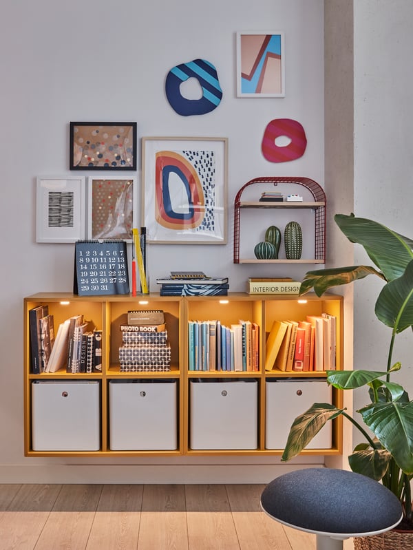 Two golden-brown cabinets put together on the wall, showing books and white boxes. Above are framed colourful illustrations.