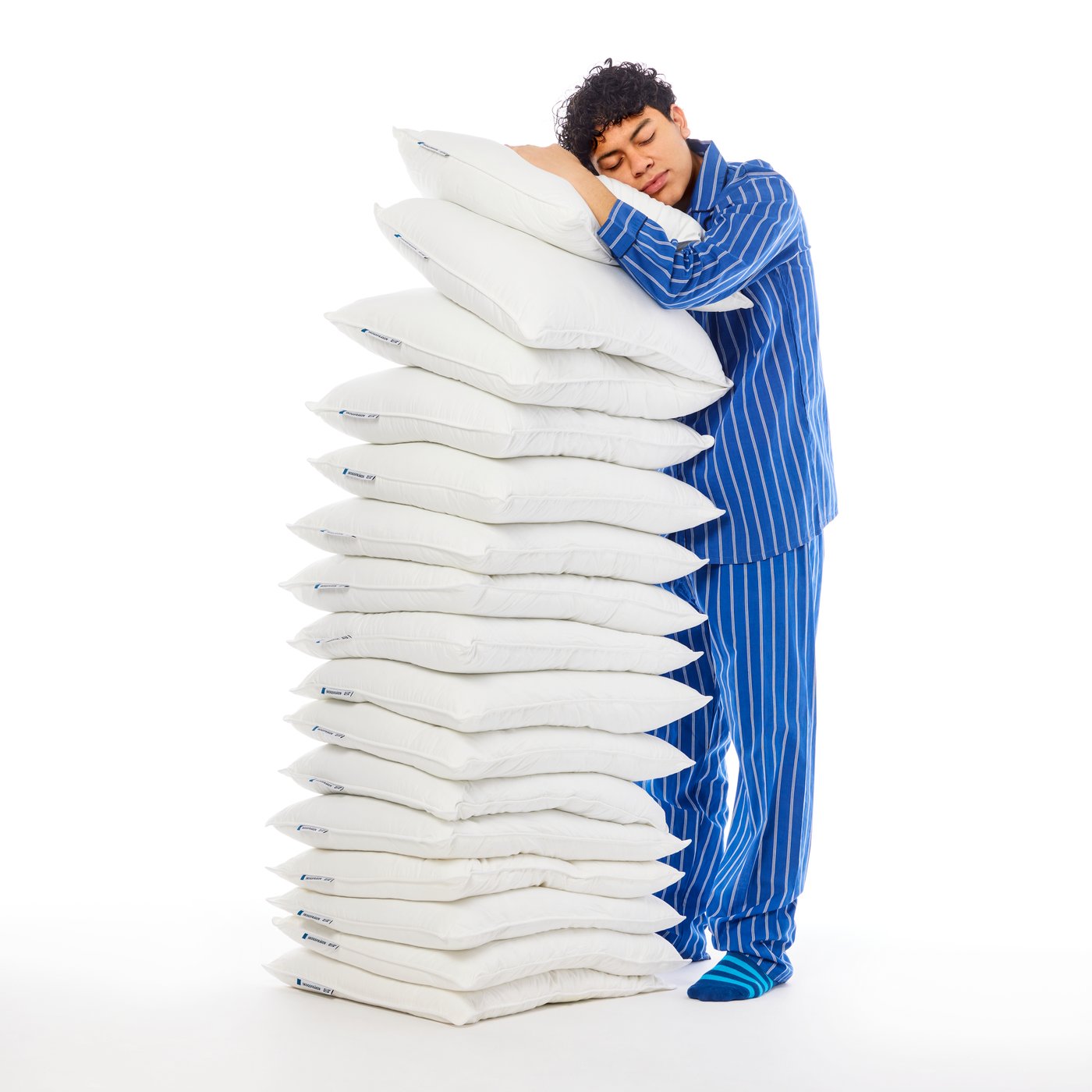 A person in blue striped pyjamas stands leaning forward into a stack of white SKOGSFRÄKEN pillows in a white space.