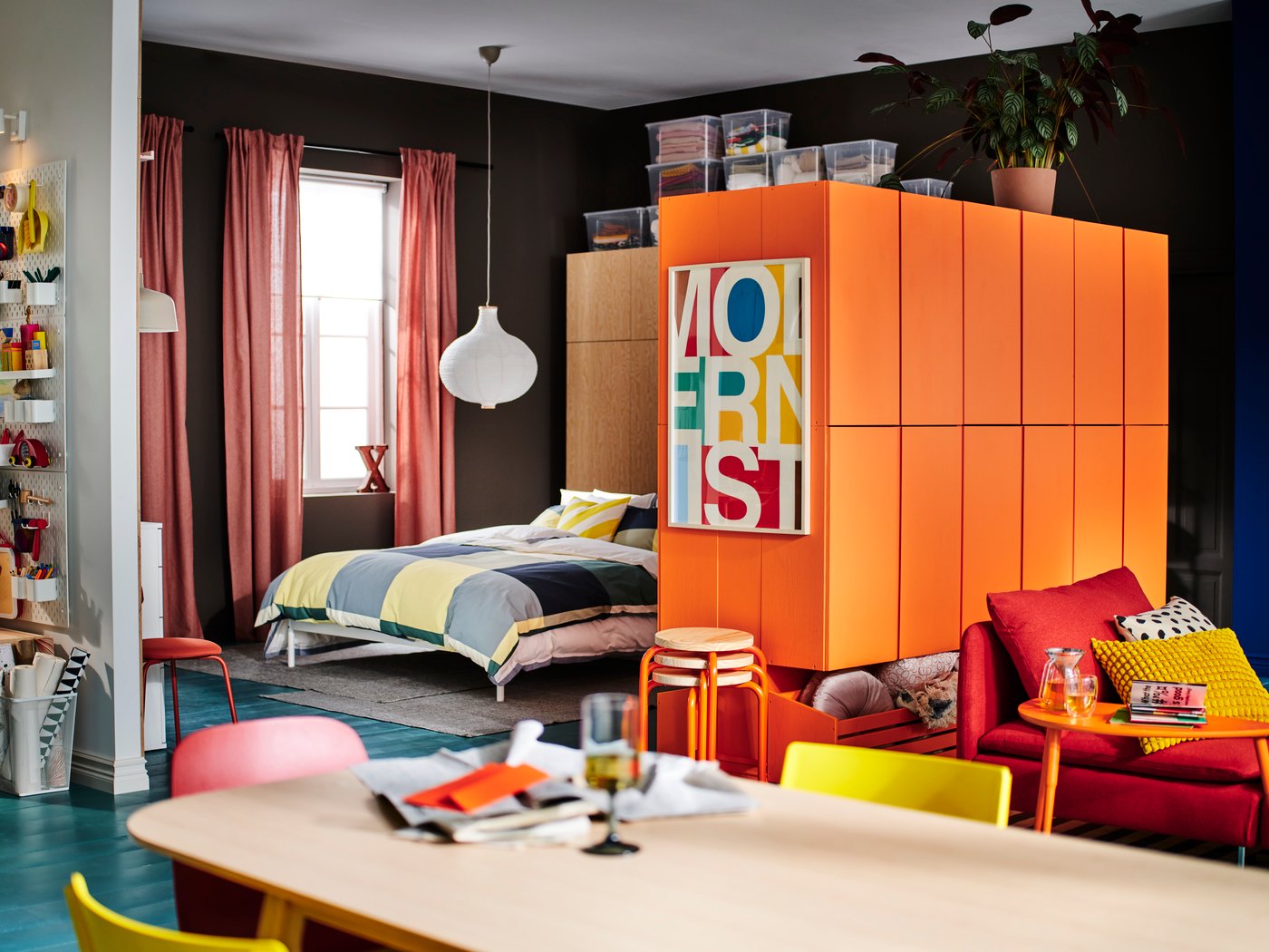 Es ist ein heller Raum der sehr bunt eingerichtet wurde. Neben einem Bett steht ein orangener Kasten mit einem bunten Bild auf der Seite. Auf dem Bett liegt blau-gelbe Bettwäsche und am Boden liegt ein baluer Teppich.