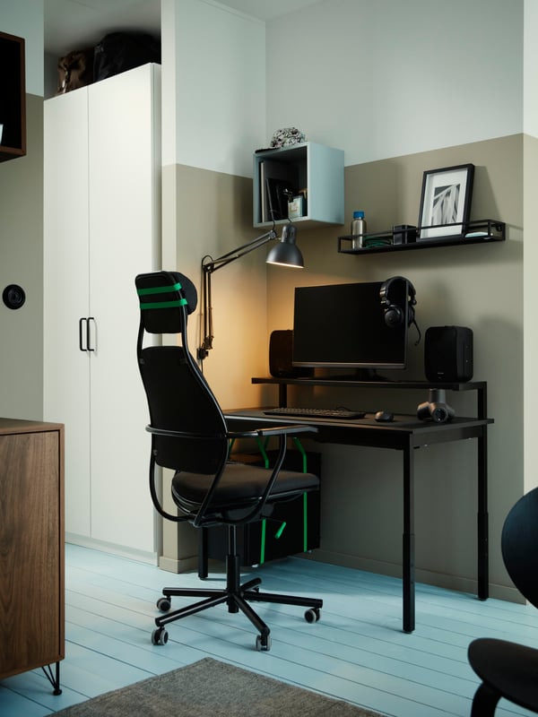 A LÖPARBANA gaming chair stands at an UTVISNING gaming desk with shelf in a corner with some storage mounted on a wall.