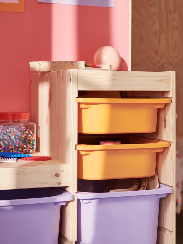 A light white stained pine TROFAST frame holds light orange and lilac boxes filled with art materials and toys.
